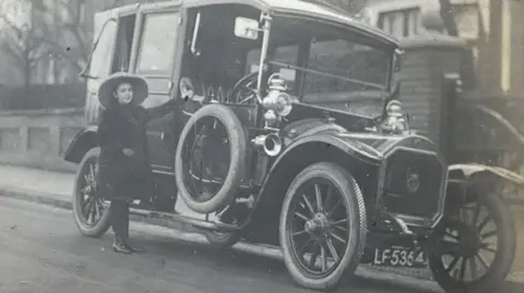 Michelle Bullivant A black and white mage showing Elsa beside a car on Woodborough Road, Putney, London. She is wearing a broad brim hat, a black knee-length coat over black tights and shoes. Her left hand is holding the window rim of the car on the driver's side and she is smiling.