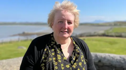 Sheena MacKellar stands with her back to a stone wall - beyond which is a stretch of grass and an island coastline. Sheena has short blonde hair and is wearing a black and yellow sunflower pattern blouse and a black cardigan