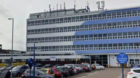 A large, five-storey building with a grey and blue front and a large car park in the foreground