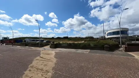 Google In the foreground is a wide path with a trail of sand lying across it. There is a short concrete wall and street lamps running alongside it. Behind is a grassy area which has some parked cars and a bus to the left. On the right, there is a modern, white, curved building with large glass windows. The day looks bright with white clouds scattered across a blue sky.
