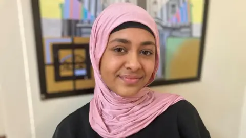 Fatima, with a pink headscarf on, standing in front of a wall with artwork on, she has a black top on and is smiling to the camera. 