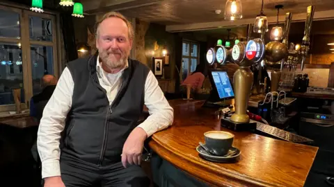 A councillor called David Gray sitting at a bar in a pub. Next to him is a cup with a hot milky liquid in. Various beer brands are illuminated on the beer taps on the bar, which is made of wood. Mr Gray is wearing a white long-sleeved shirt and a grey woollen-style gilet. He has a beard and is smiling at the camera