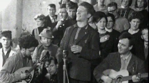 Una imagen en blanco y negro de un grupo de músicos y miembros de la audiencia de 1964. La banda se sienta al frente con la audiencia detrás de ellos. De izquierda a derecha de los miembros de la banda hay un hombre con cabello oscuro corto tocando un instrumento de cuerda, un hombre de pie con cabello corto y una chaqueta en frente de un micrófono y a su izquierda hay un hombre con cabello corto sentado mientras toca una guitarra.