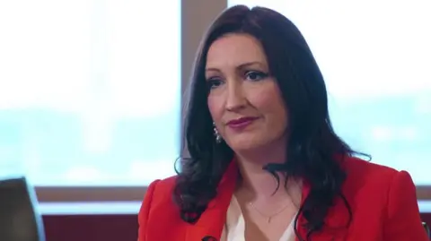 Emma Little-Pengelly is wearing a red blazer and white shirt. She has a fine necklace on and silver drop down earrings. She has long curly black hair and brown eyes.