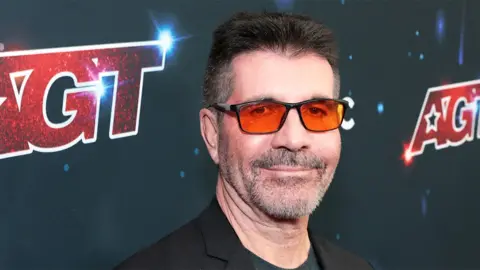 Getty Images Simon Cowell, a man wearing orange sunglasses while smiling.