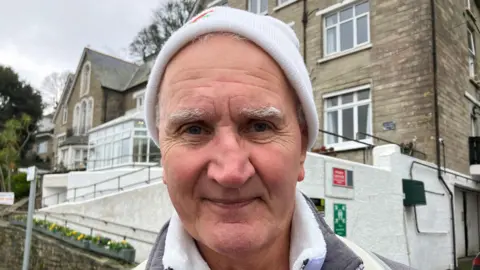 Man smiling at the camera he is wearing a woolly hat and has bushy white eyebrows