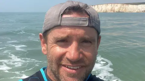 A selfie shot of Stephen Hopson wearing a backwards baseball cap with the sea and white cliffs in the background