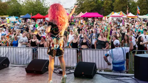 Pride in Gloucestershire A person in a dress singing to a large crowd of people on a stage