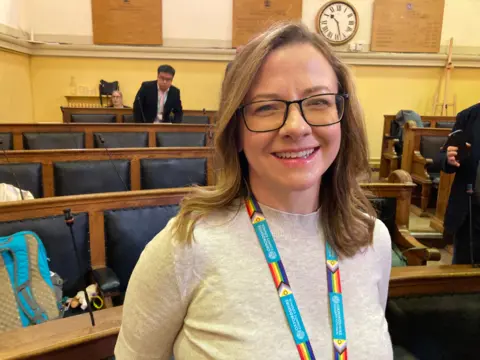 A woman is smiling at the camera. She is wearing glasses an a council lanyard. The photo is taken in a council chamber.