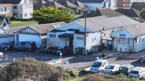 Eddie Mitchell A fire-damaged white building in a coastal English town. A burnt-out car is parked in front of the mosque entrance. There is police tape around the scene and police officers standing guard.