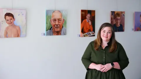 Photographs by Sara Jade Magee is a young woman with long brown hair which is lighter towards the ends. She is wearing a long-sleeved green dress with buttons up the front. She is smiling at the camera and is standing in front of some of the paintings in her collection which are hanging on a white wall. 