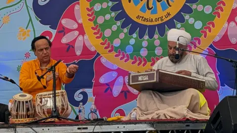 Art Asia Two men perform on stage in front of a colourful backdrop. They are both sat on the ground playing instruments. The man on the left is playing the tabla drums while the other is playing the santoor.