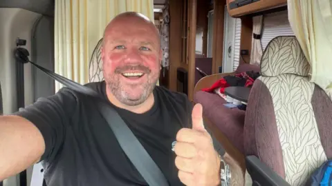 Russ Lee A selfie of Russ Lee smiling and giving a thumbs up to the camera while sitting in passenger seat of his camper van wearing a black t-shirt. 