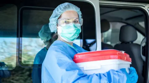 a female medic wearing blue scrubs and a face mask holds an organ transplant container, which is a white plastic tub with a red lid. She appears to be stood to the side of a vehicle with a sliding door