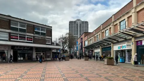 Empty town centre with shops and people walking by the shops. An industrial looking office block looms over the scene.