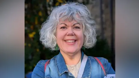 Maxine Webb Maxine Webb is smiling while wearing a denim jacket on top of a cream top. She has curly hair and is standing outside.