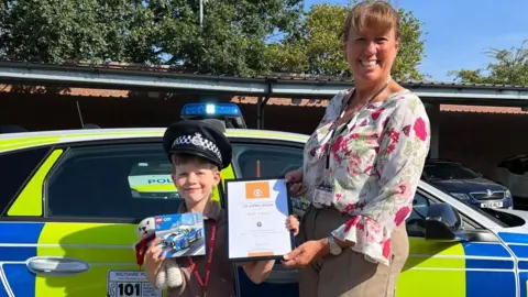 Wiltshire Police Eli is stood in front of a police car next to Deb Smith, a woman with ginger hair and a flowery top. They are both holding a certificate and Eli is holding a lego set and wearing a police hat. 