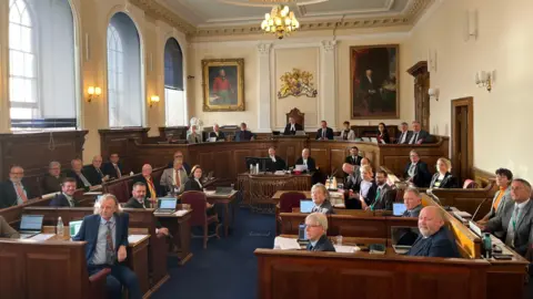 BBC A group of people wearing suits look towards the camera in a courtroom setting. 