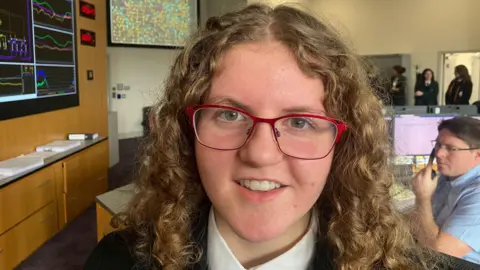 Eva with light brown curly hair and red glasses. She is wearing a blazer, white shirt and tie. There is a classroom behind her.