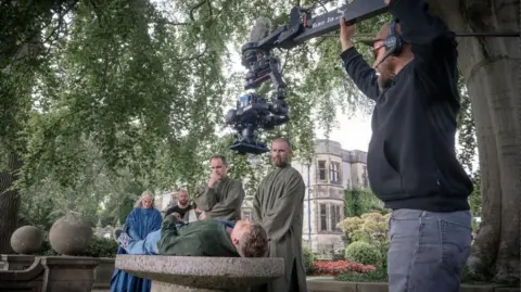 Run Away An actor is lying on a concrete slab surrounded by other actors. A cameraman has positioned the filming equipment above him. 