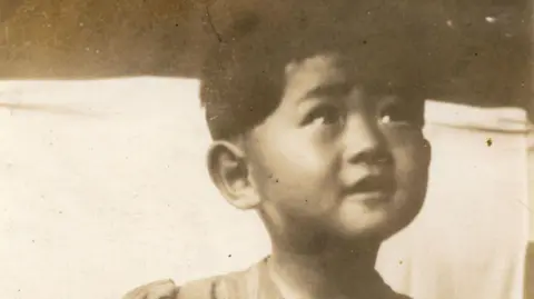 BBC/Minnow Films/Michiko Kodama Michiko Kodama pictured as a small child in a black and white photograph. She is not looking at the camera, but looking up at someone with a happy expression on her face.