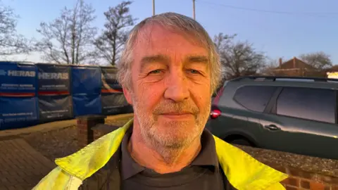 John Devine/BBC Mark Jackson looking into the camera on a driveway. He has short grey hair and stubble and is wearing a hi-vis jacket and black jumper.