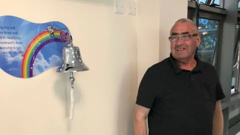 Peter Firth in a black top with short hair and glasses looking towards a silver bell on a beige wall. The bell is mounted on a poster with a rainbow and sky scene with a positive message. The bell has a rope on it.