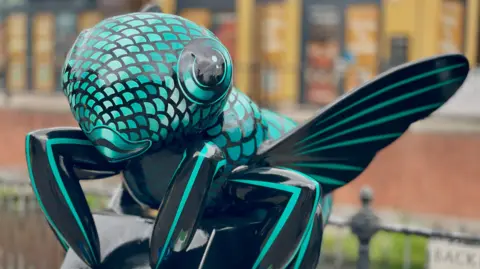 A close-up of a bee sculpture painted in black and turquoise with a fish scale pattern on its body.