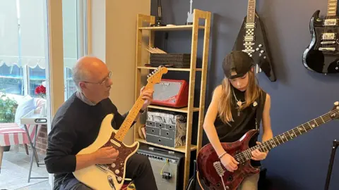 Lee sits in a blue-walled room with guitars on the wall and the doors open to a conservatory in the background. He is wearing a dark grey jumper and is playing a cream-coloured electric guitar, while looking at his grandson. Olly is standing next to him playing a dark brown electric guitar. The boy is wearing a back-to -front baseball cap and a black vest top with grey jeans. He is also wearing a beaded necklace. 