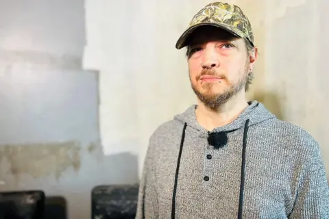 BBC Gary Bennett, a man wearing a baseball cap and grey hooded top, in front of mouldy furniture in his council flat. 