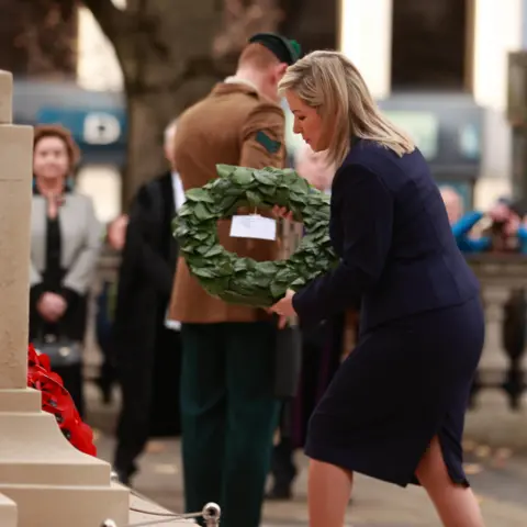 PA Media Michelle O'Neill, a woman in a dark suit, is placing a green wreath at a stone memorial. A soldier stands behind her, and a small crowd is watching quietly in the background.