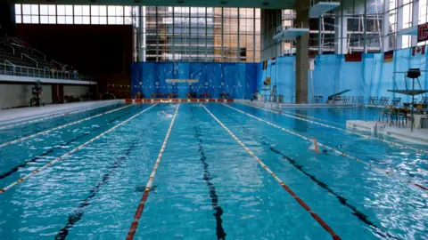 A swimming pool with lane markers seen from one end, with tiered seating to th left. Windows are covered in blue sheeting.