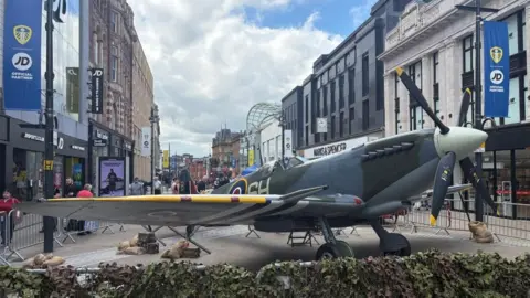 A military aircraft display set up in the middle of Briggate in Leeds during a public event. The plane is positioned diagonally across the street behind metal crowd-control barriers and painted in camouflage tones of dark green and grey.