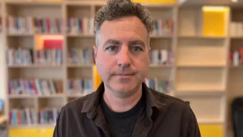 Tim Evans poses for a photo in a library room at his organisation's office in Hereford. Behind him is shelving with books on. He is wearing a black t-shirt and brown shirt over the top.
