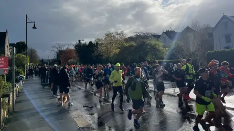 Hundreds of runners running along a road. They are all dressed in sports gear. Trees and houses line the side of the road. 
