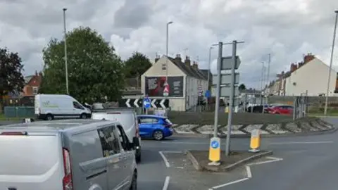 A busy roundabout with a lot of cars and vans. A suburban street is visible in the distance.  