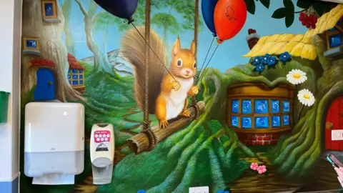 A mural painted on a hospital wall depicting a squirrel on a wood log swing and a small cottage embedded in the ground surrounded by small tree homes.