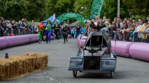 A batman go-kart travelling along a closed road as part of a race.  Crowds are gathered at the side of the road. The crowd is separated from those involved in the race by metal barriers with hay bales in front of them.