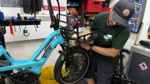 A man with a cap holds up a large blue bike to the camera. A yellow bucket and various tools and equipment can be seen behind him.