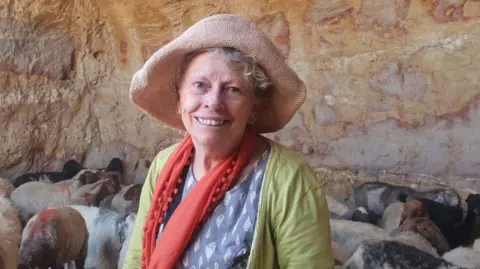 International Solidarity Movement A woman smiling at the camera wearing a hat, red scarf, green jumper and a blue top. You can see a group of sheep behind her and she appears to be in a cave.
