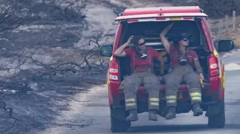 Two fire officers, one female, are in the back of a red fire vehicle with the boot door open. They are wearing fire safety clothing and around their necks are breathing masks. Both wear sunglasses. Around them are the remnants of burnt trees and grassland. The red vehicle appears to be driving on a road away from the camera.