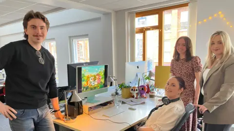 Three woman and a man standing around a desk in an office. 