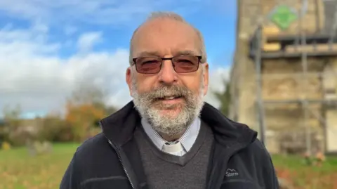 A middle-aged man with cropped light-grey hair and a full beard stands in front of a rural parish church set in green grounds and under a blue sky. He is wearing tinted rectangular glasses, a black fleece, grey jumper, blue and white striped shirt and white dog collar. 
