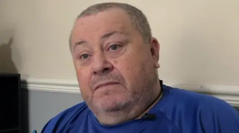 BBC A man looks to the left of the camera. He is wearing a blue T shirt and has short grey hair and stubble. He is sitting against a plain wall.