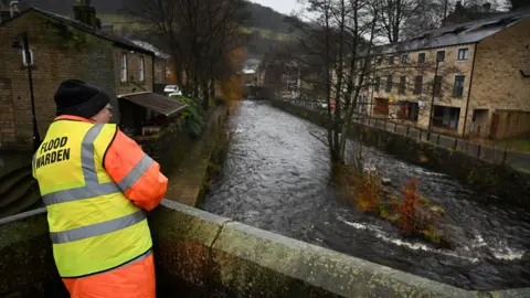 A man wearing a luminous jacket with the words 'flood warden' written on the back observes a river from a bridge above. Houses sit either side of the river and a leafless tree sits in the middle of the water itself.