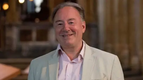 Elgar Festival 2025 Ian Venables with mainly dark hair is smiling at the camera. He is wearing a light jacket.