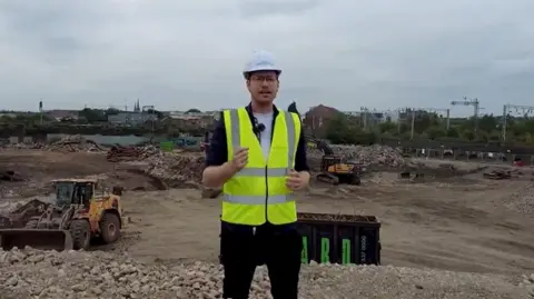 A still from a video shows a man in a white hard hat and hi-viz vest talking to the camera against a backdrop of rubble  with two yellow diggers. In the background, overhead electric power lines run along a railway line. 