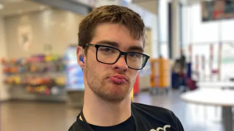 A 21 year old man with short dark hair and a fringe. He has black glasses and a black t-shirt. He wears a hearing aid.