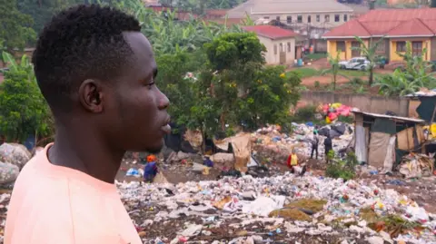 Pangeran BBC Okuku dengan kaus merah muda yang menghadap ke tempat sampah Kiteezi di Kampala, Uganda, dengan rumah-rumah terlihat di latar belakang.