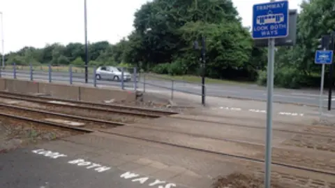 A crossing on a tramway. There are two poles with blue signs reading 'Tramway look both ways' and white paint on the road which says 'Look both ways'.
